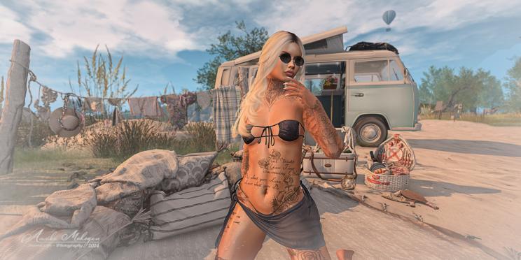 Das Bild zeigt eine aufwändig tätowierte Annika in einem schwarzen Bikini mit Sarong kniend vor einem Wohnmobil am Strand